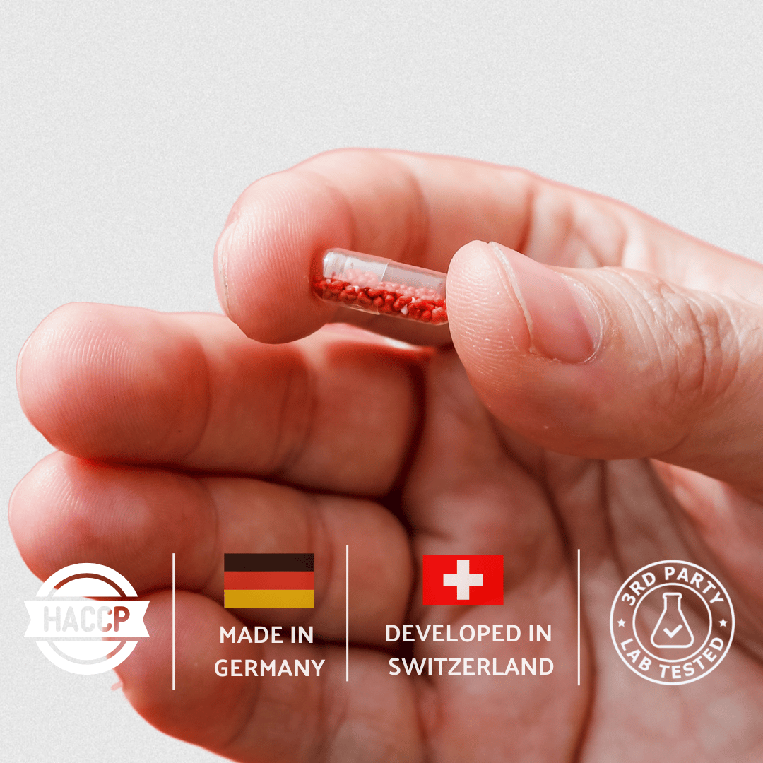 Hand holding a small red capsule with certification logos on a light gray background