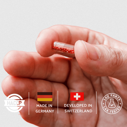 Hand holding a small red capsule with certification logos on a light gray background